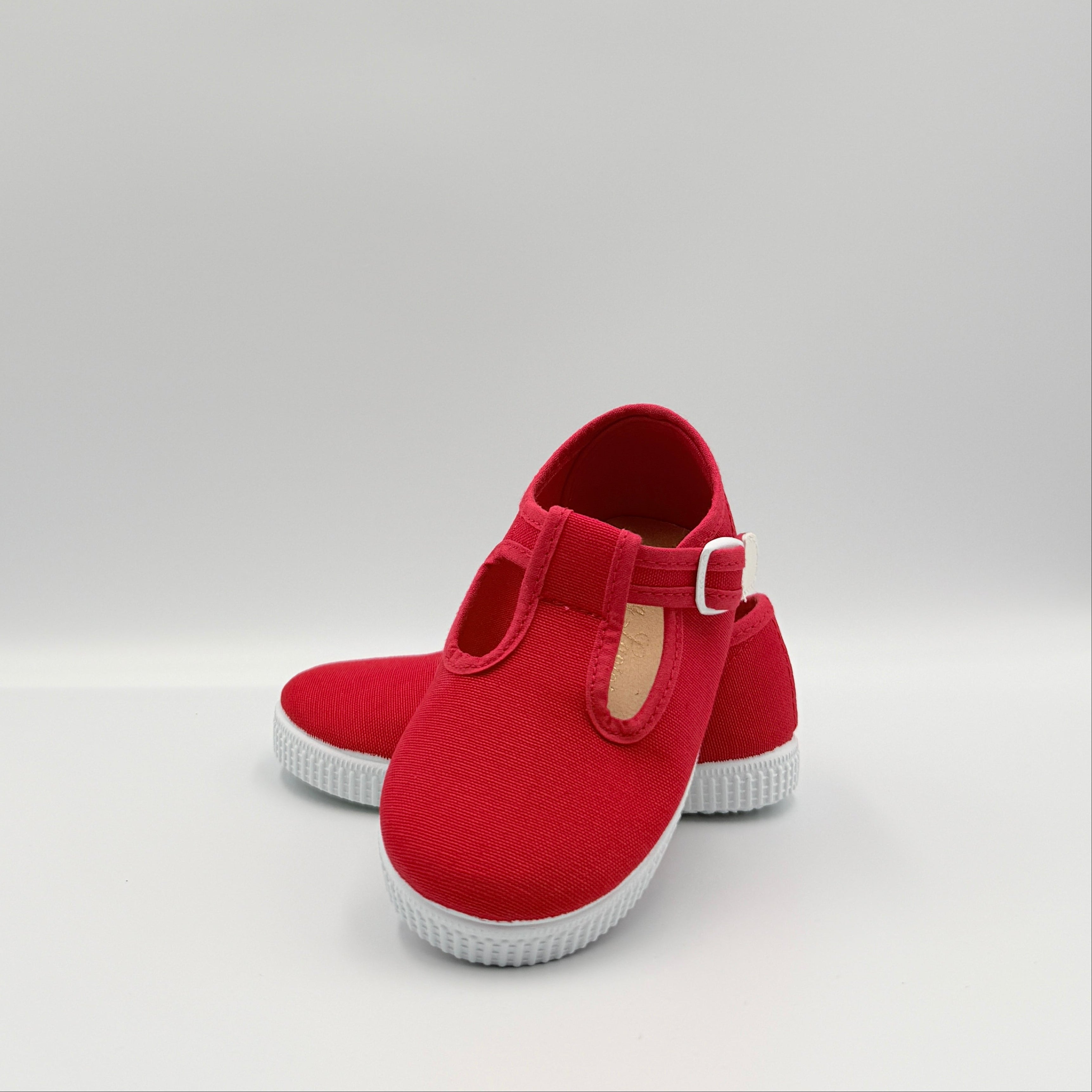 A pair of red canvas Mary Jane-style shoes with white soles and a small beige pull tab, photographed on a light gray studio background.