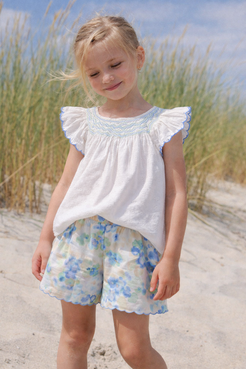 Young girl in a white top with ruffled sleeves and floral shorts standing on a sandy beach.