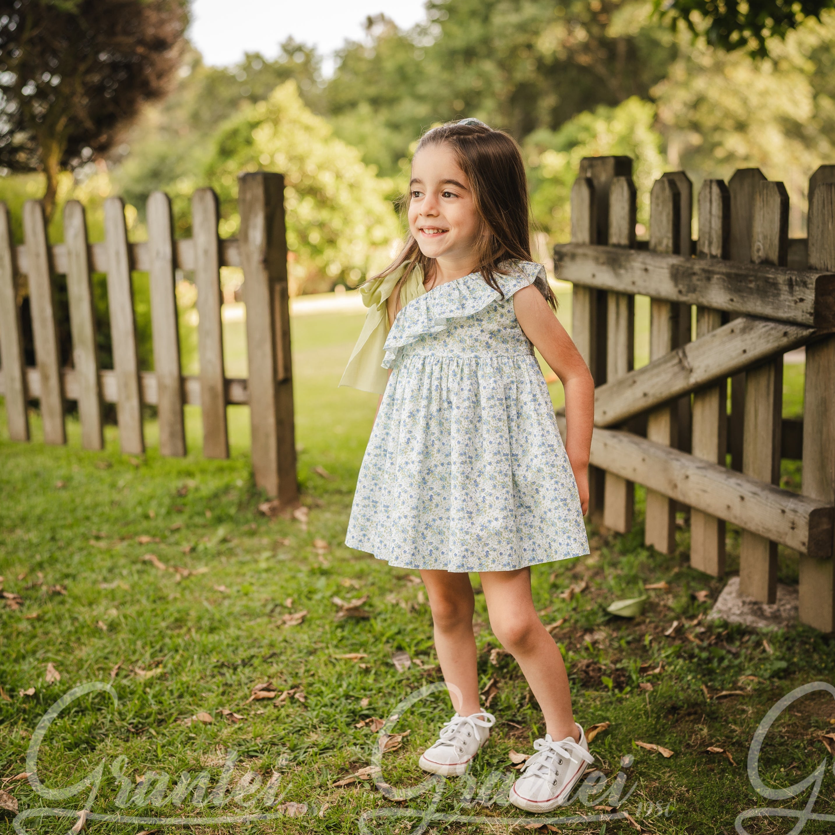 Young girl in a floral dress standing in a grassy area with a wooden fence in the background.
