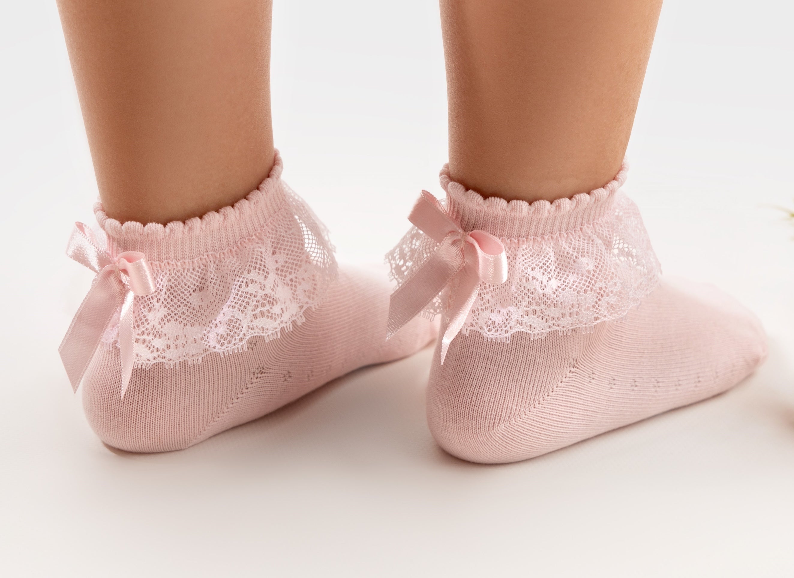 A close-up of a person's feet wearing light pink ankle socks with white lace trim and ribbon bows, standing on a white surface next to pink roses.