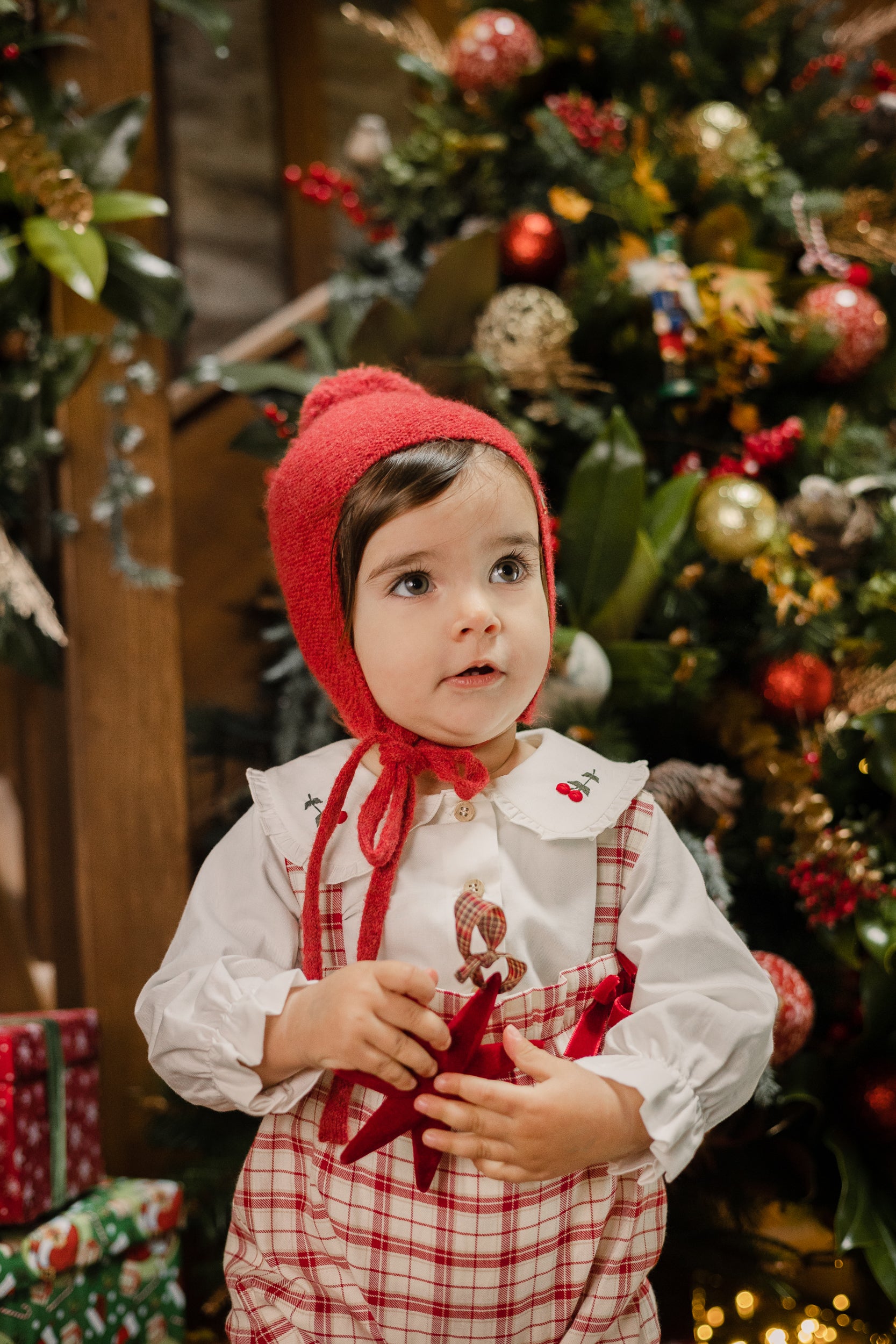 Child wearing a red bonnet and plaid outfit in front of a decorated Christmas tree.