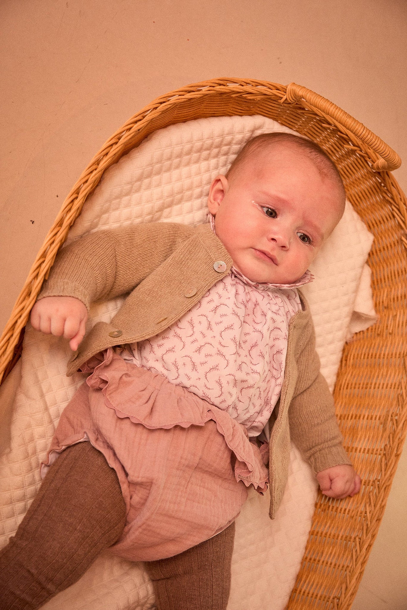 Baby in a wicker crib wearing a brown coat and pink dress.